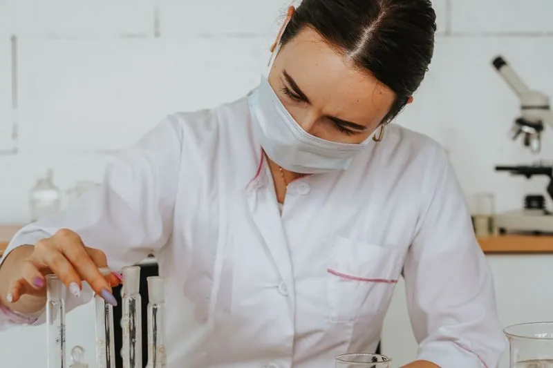 Chercheuse en blouse blanche et masque chirurgical manipulant des tubes à essai, un microscope en arrière-plan