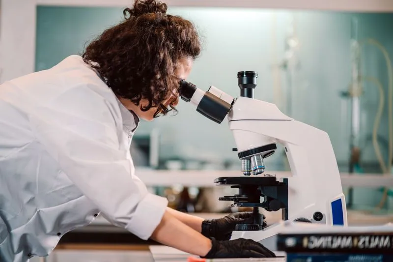 Chercheuse aux cheveux bouclés en blouse blanche et gants noirs, penchée sur l'oculaire d'un microscope