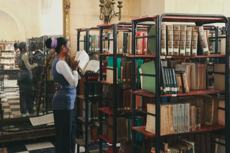 Femme feuilletant un livre ancien devant des étagères métalliques chargées de volumes reliés, dans une bibliothèque