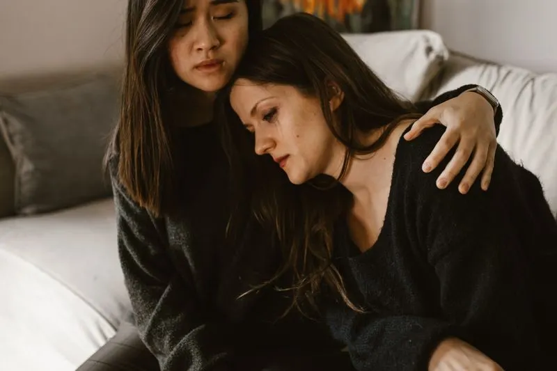 Femme en larmes, la tête posée sur l'épaule d'une amie qui la serre contre elle sur un canapé