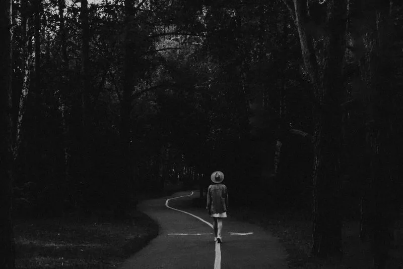 Femme au chapeau marchant seule sur une allée sinueuse à travers une forêt sombre, vue de dos, en noir et blanc