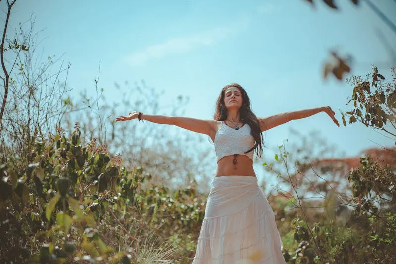 Femme aux bras grands ouverts et aux yeux fermés, debout au milieu de la végétation sous un ciel bleu