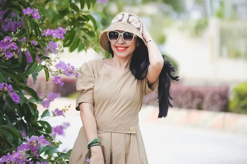 Femme souriante en robe beige, bob en crochet et lunettes de soleil, devant des fleurs mauves