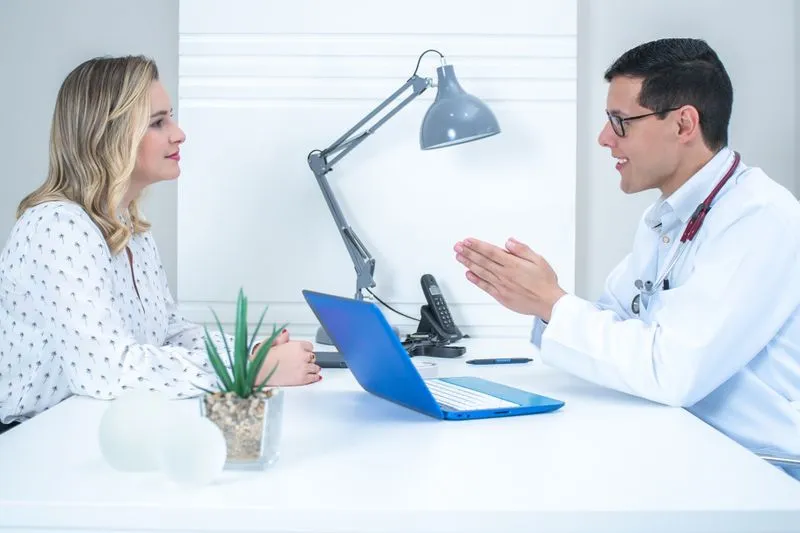 Médecin en blouse blanche échangeant avec une patiente assise face à lui, de l'autre côté de son bureau
