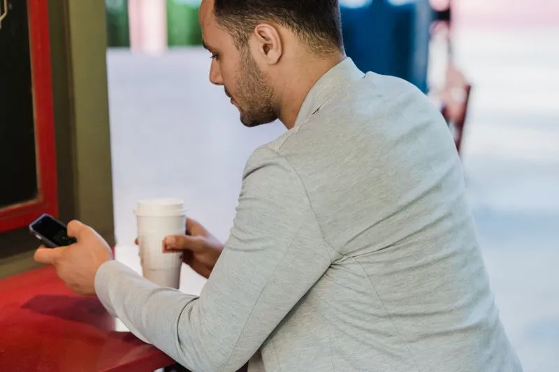 Homme attablé à une terrasse de café, téléphone dans une main et gobelet de café dans l'autre