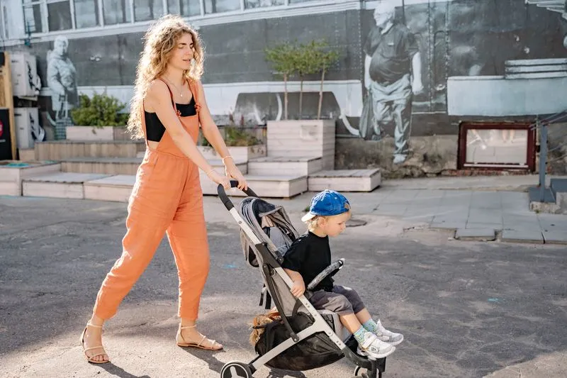 Jeune femme en combinaison orange qui pousse une poussette où est assis un petit garçon, dans une rue ensoleillée