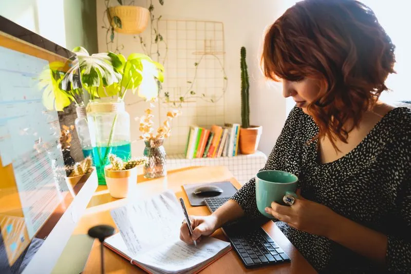 Femme aux cheveux auburn qui écrit dans un carnet à son bureau, une tasse verte à la main
