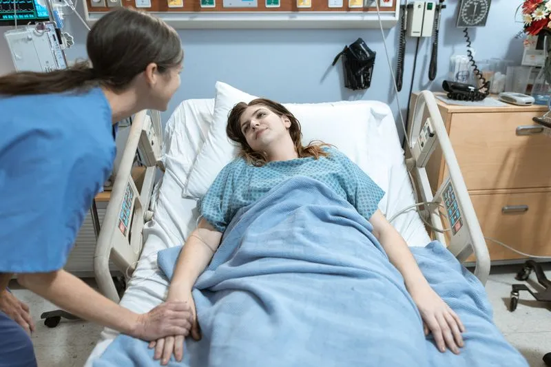 Soignante en blouse bleue qui rassure une jeune femme allongée dans un lit d'hôpital