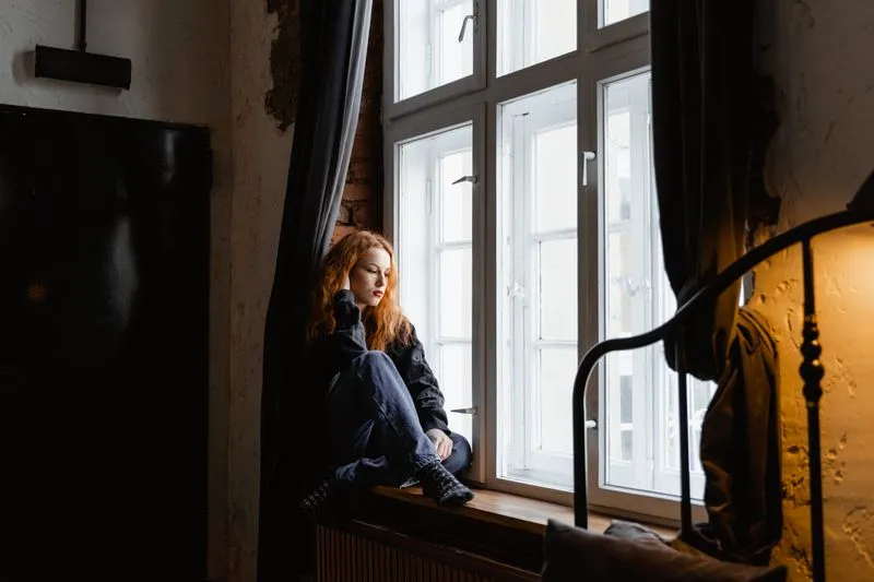 Femme assise seule sur le rebord d'une fenêtre, genoux repliés, le regard absent