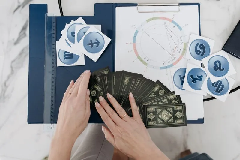 Mains étalant un jeu de tarot devant un thème astral imprimé sur un porte-bloc, entouré de cartes des signes du zodiaque