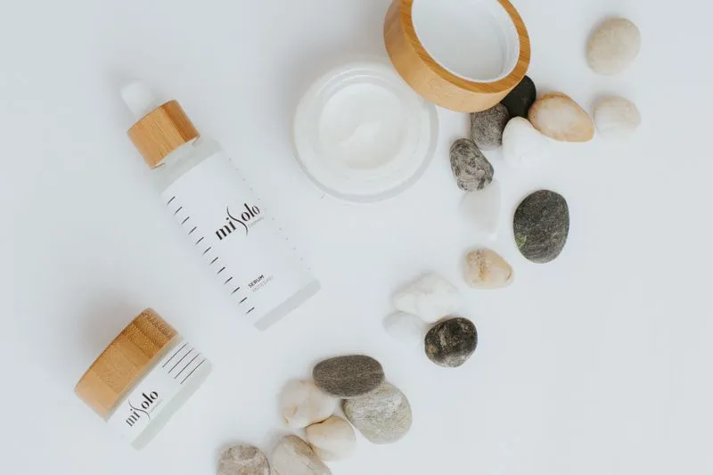 Flacon de sérum à pipette et pot de crème blanche ouvert, couvercle en bambou, posés parmi des galets sur fond blanc