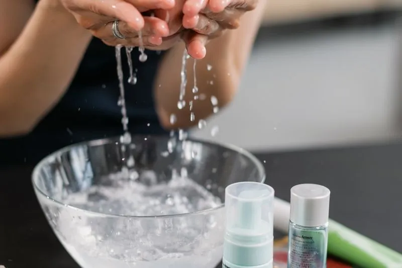 Mains qui s'aspergent d'eau au-dessus d'un bol en verre, deux petits flacons de soin posés à côté