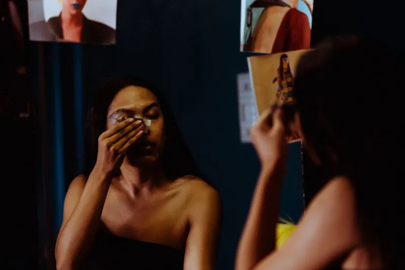 Jeune femme qui se démaquille les yeux avec un coton devant un miroir, dans une pièce sombre