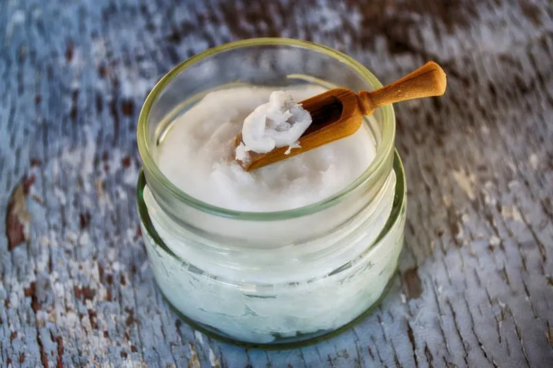 Pot en verre rempli d'huile de coco solide, avec une petite cuillère en bois posée dessus, sur une planche en bois patinée