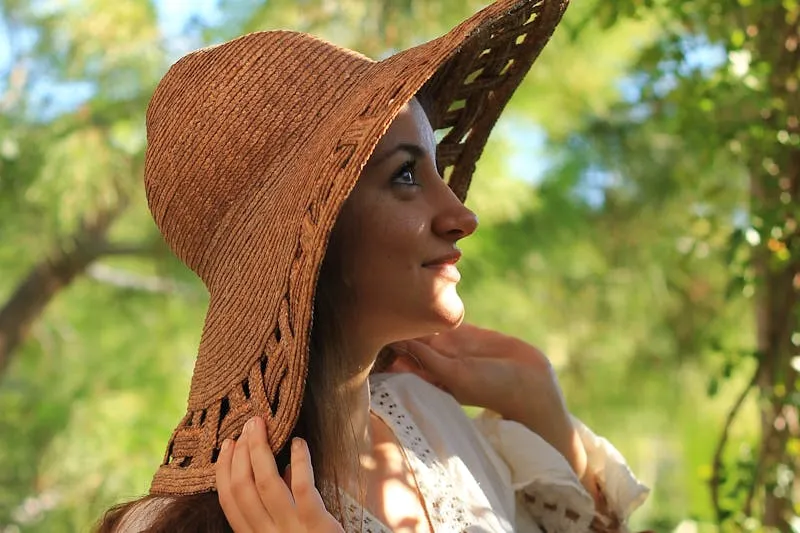 Jeune femme en chapeau de paille à larges bords, le visage au soleil sous des arbres
