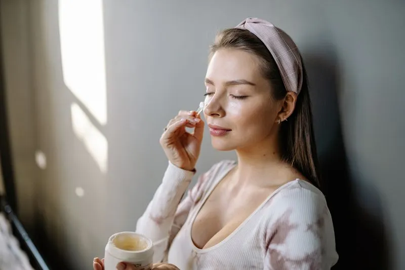 Jeune femme les yeux fermés, bandeau rose à carreaux, prélevant de la crème dans un pot avec une petite spatule près de son visage, dans la lumière d'une fenêtre