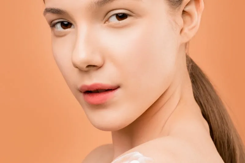 Jeune femme aux cheveux attachés, regard par-dessus l'épaule, une noisette de crème sur l'épaule, fond orangé