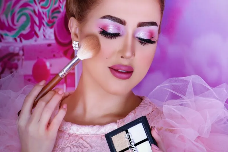 Femme au maquillage de soirée rose qui passe un grand pinceau à poudre sur le haut de la pommette, une palette de poudres claires à la main