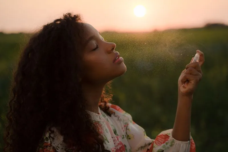 Jeune femme aux cheveux bouclés qui vaporise une brume près de son visage, les yeux fermés, dans un champ au coucher du soleil