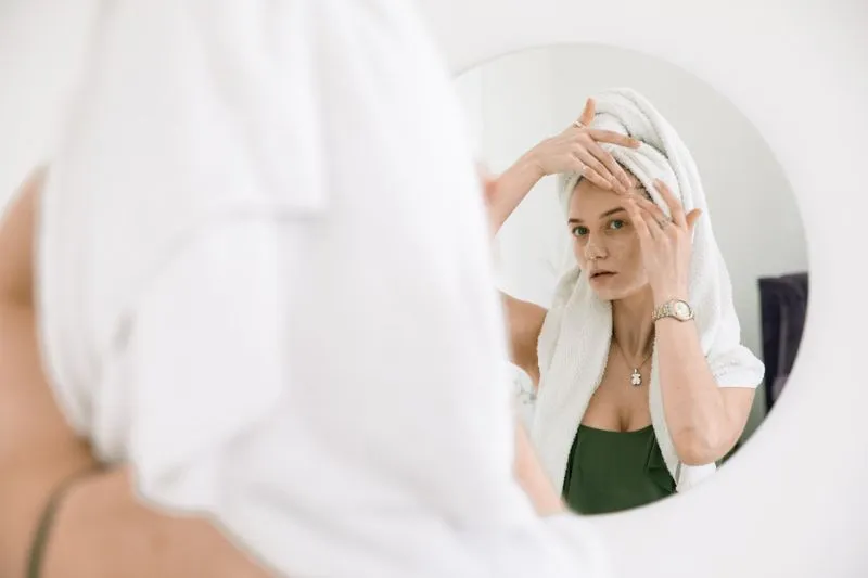 Femme aux cheveux enroulés dans une serviette qui fait pénétrer sa crème du bout des doigts, face à un miroir rond