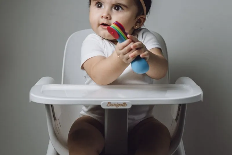 Petite fille assise bien droite dans sa chaise haute, qui tient un jouet à deux mains près de sa bouche