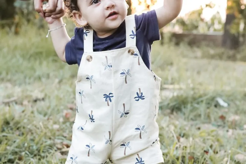 Bébé en salopette qui marche dans l'herbe, les bras levés, tenu par un adulte