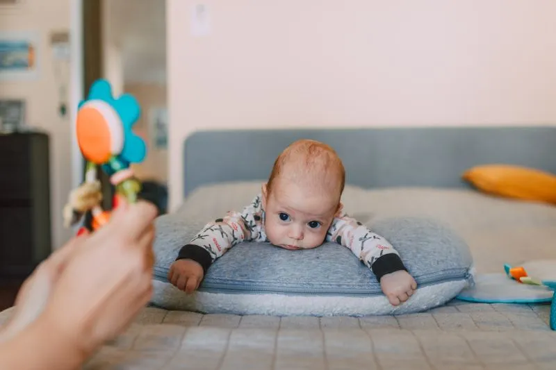 Bébé à plat ventre sur un coussin, le menton posé, qui fixe un hochet tendu par un adulte