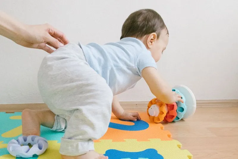Bébé à quatre pattes sur un tapis en mousse, qui tend la main vers un jouet, un adulte la main posée sur son dos