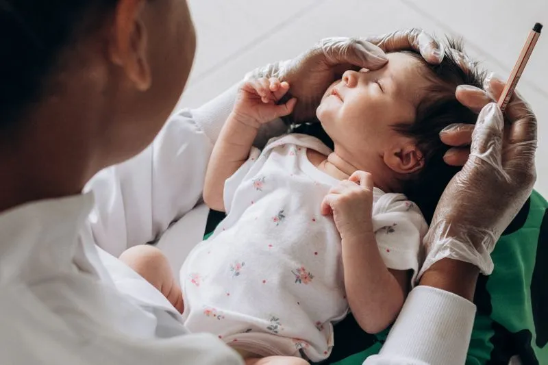 Une professionnelle de santé en blouse blanche, mains gantées, examine la tête d'un nourrisson aux yeux fermés, en body blanc à petites fleurs