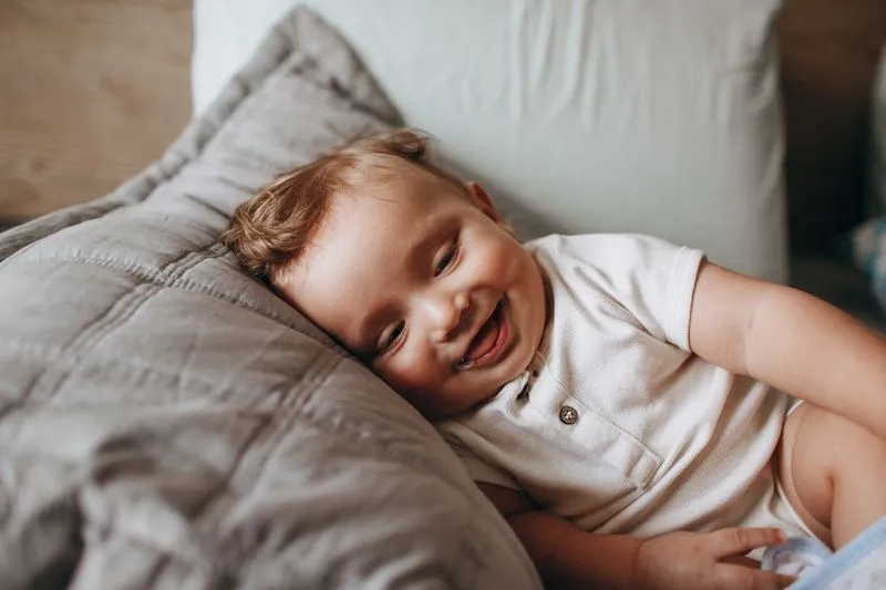 Bébé en body blanc qui rit aux éclats, la tête posée sur un coussin gris