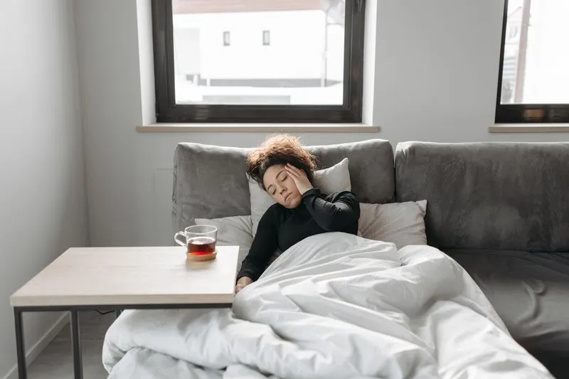 Femme allongée sur un canapé sous une couette, la main sur la tempe, une tasse de thé posée à côté