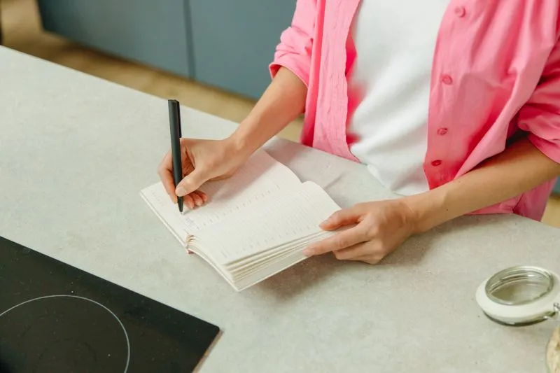 Femme en chemise rose qui écrit dans un carnet ouvert posé sur un plan de travail