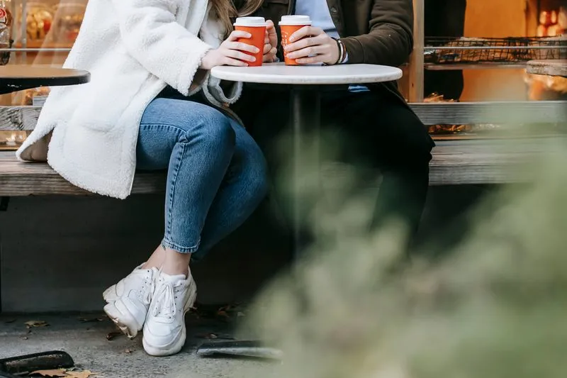Un couple assis côte à côte sur un banc devant un café, chacun tenant un gobelet orange posé sur une petite table ronde