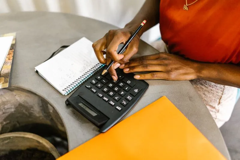 Mains d'une femme tapant sur une calculatrice, un crayon à la main, à côté d'un carnet de notes et d'une pochette orange