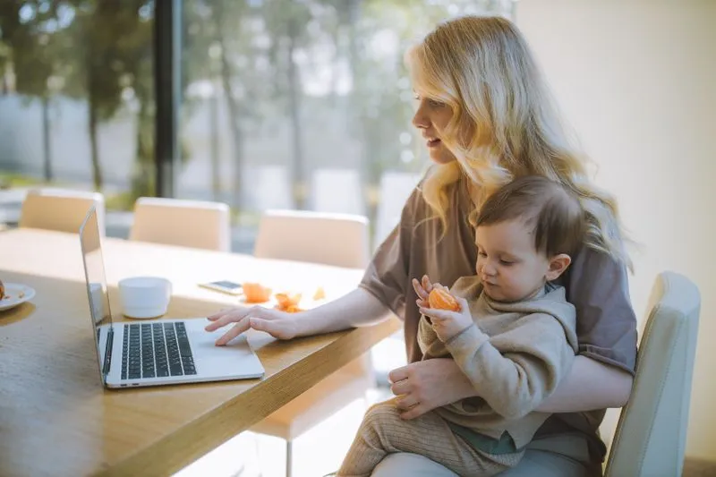 Mère travaillant sur un ordinateur portable à une table de salle à manger, son bébé sur les genoux, un fruit à la main
