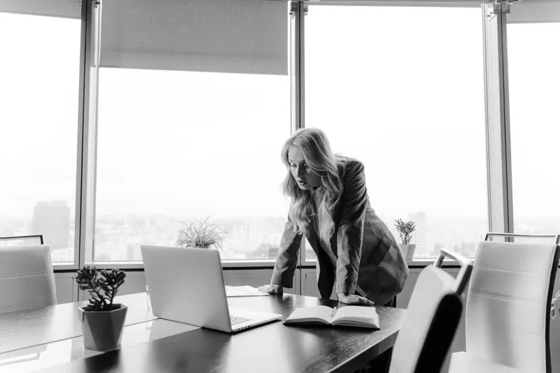 Femme en blazer penchée sur une table de réunion, devant les baies vitrées d'un bureau en étage qui domine la ville, en noir et blanc