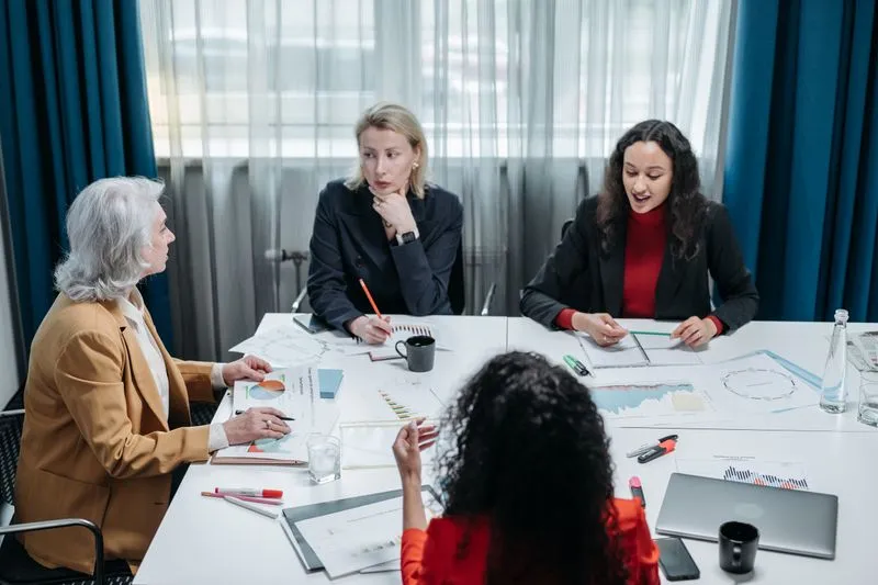 Quatre femmes en réunion autour d'une table blanche couverte de graphiques imprimés, de carnets et d'un ordinateur portable