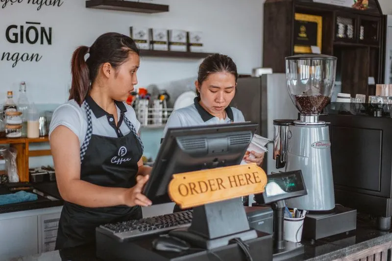 Deux employées en tablier derrière le comptoir d'un café, l'une à la caisse, sous un panneau « Order here »