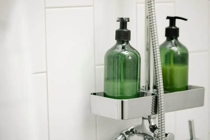 Deux flacons pompe verts sur la tablette chromée d'une douche, à côté du flexible
