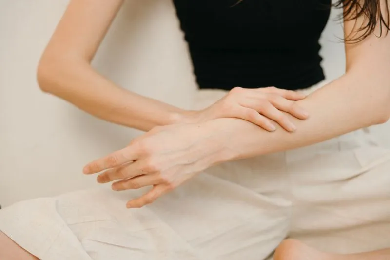 Femme en débardeur noir et pantalon de lin beige qui masse son avant-bras du plat de la main