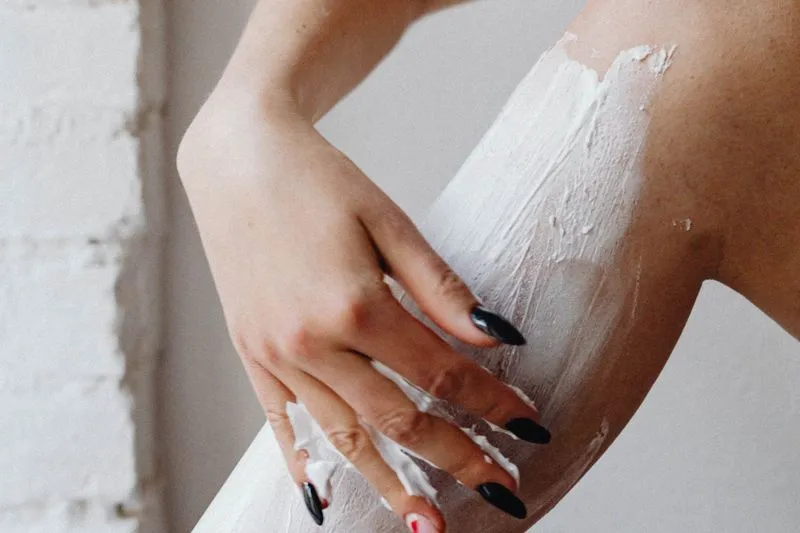 Une main aux ongles vernis de noir étale une épaisse couche de crème blanche sur une jambe, devant un mur de briques blanches