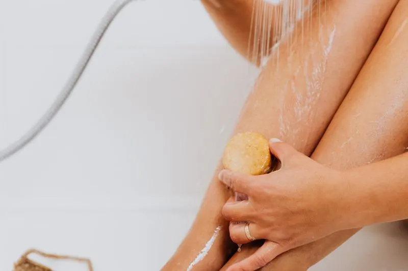 Une femme frotte sa jambe avec un pain de gommage sous le jet de la douche