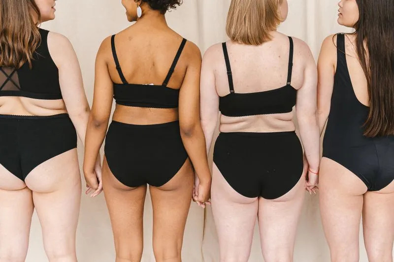 Quatre femmes de dos en maillot et sous-vêtements noirs, aux silhouettes et aux carnations différentes, se tenant la main