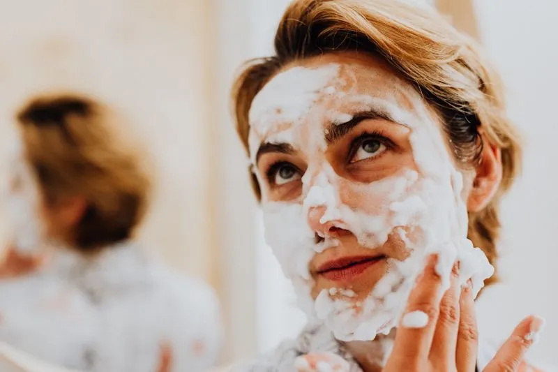 Une femme au visage couvert de mousse nettoyante la masse du bout des doigts devant un miroir