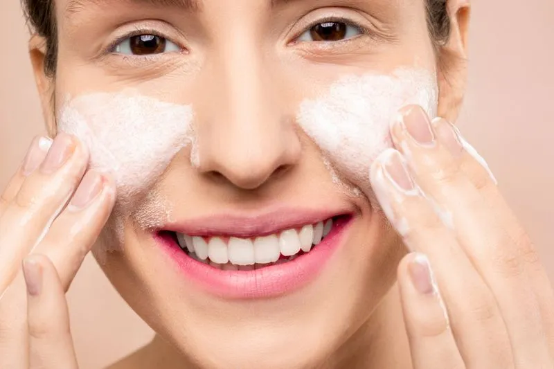 Une femme souriante étale une mousse nettoyante blanche sur ses joues du bout des doigts