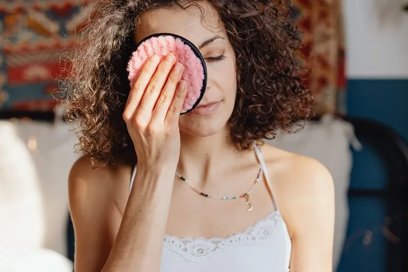 Une femme aux cheveux bouclés tient un disque démaquillant lavable rose posé sur son œil fermé