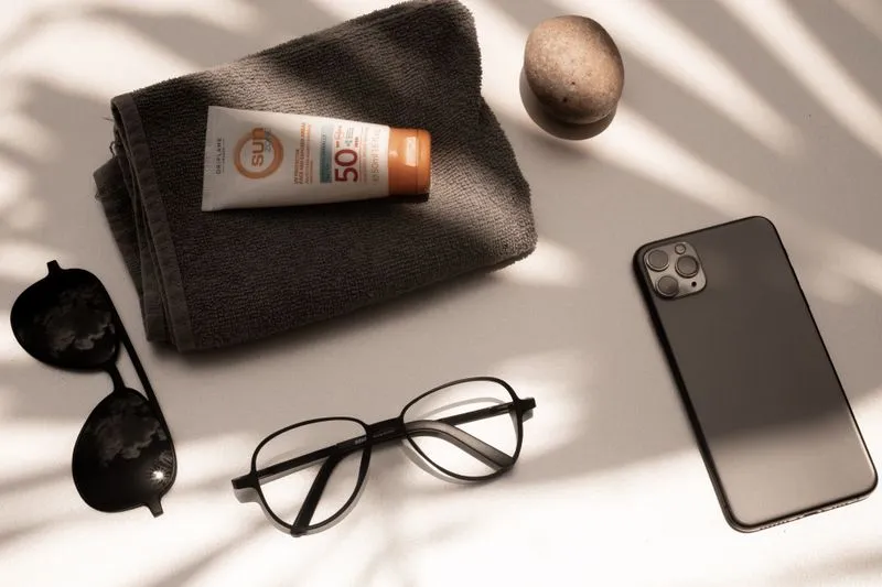 Tube de crème solaire SPF 50 sur une serviette, lunettes de soleil, lunettes de vue, galet et téléphone posés à côté, dans l'ombre de palmes