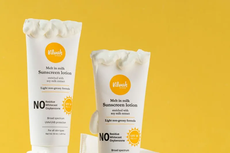Deux tubes de crème solaire dont la texture crémeuse déborde du bouchon, sur fond jaune