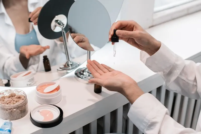 Femme qui fait tomber quelques gouttes dans sa paume devant un miroir de coiffeuse, entre des pots de soin et un petit flacon brun