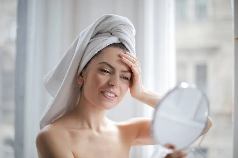 Femme aux cheveux enroulés dans une serviette blanche qui observe sa peau dans un miroir à main